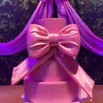 Bolo decorativo rosa de dois andares com laço de cetim em mesa de aniversário infantil.