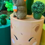 Capivara de pelúcia com chapéu de aniversário sobre cilindro decorativo com rosto de capivara.
