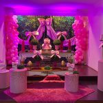 Cenário completo de festa infantil rosa com balões, laços de cetim, mesas e cilindros decorativos.