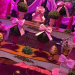 Mesa de aniversário infantil com bolo rosa, laços de cetim e arranjo ornamental.