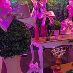 Painel de festa infantil com tecidos rosa, grandes laços de cetim, balões e bolo decorativo.